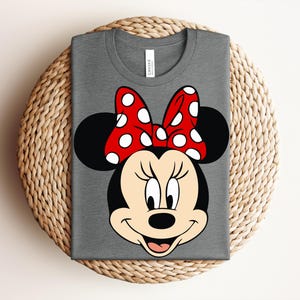 May include: A gray t-shirt featuring a graphic of Minnie Mouse's face. The design includes her signature black ears, a red bow with white polka dots, and a smiling face. The shirt is folded on a woven surface.