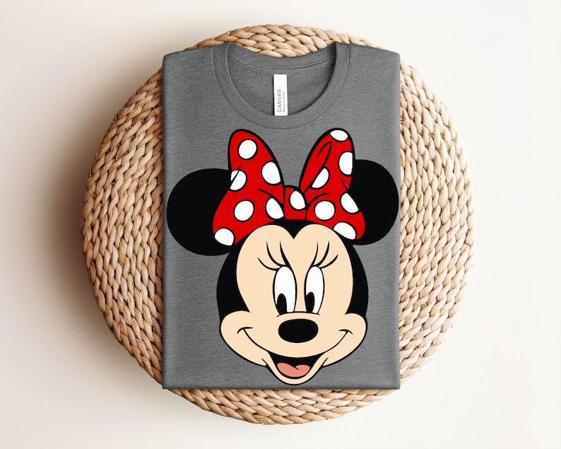 May include: A gray t-shirt featuring a graphic of Minnie Mouse's face. The design includes her signature black ears, a red bow with white polka dots, and a smiling face. The shirt is folded on a woven surface.