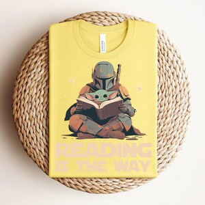 May include: A yellow t-shirt featuring a graphic of a helmeted character in armor sitting cross-legged and holding an open book with a small green creature. The text "READING IS THE WAY" is printed in large, light-colored capital letters below the graphic.
