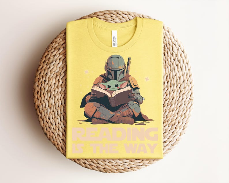 May include: A yellow t-shirt featuring a graphic of a helmeted character in armor sitting cross-legged and holding an open book with a small green creature. The text "READING IS THE WAY" is printed in large, light-colored capital letters below the graphic.