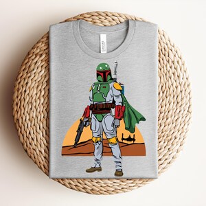 May include: A gray t-shirt featuring a graphic of a Star Wars character, Boba Fett, in his signature green and gray armor, holding a blaster. The design includes a desert landscape and a spaceship silhouette.
