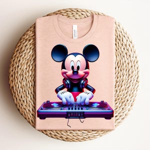 May include: A light peach-colored t-shirt featuring a graphic of Mickey Mouse as a DJ. Mickey is wearing headphones and has his hands on a purple and blue turntable. The shirt is folded on a woven, circular placemat.