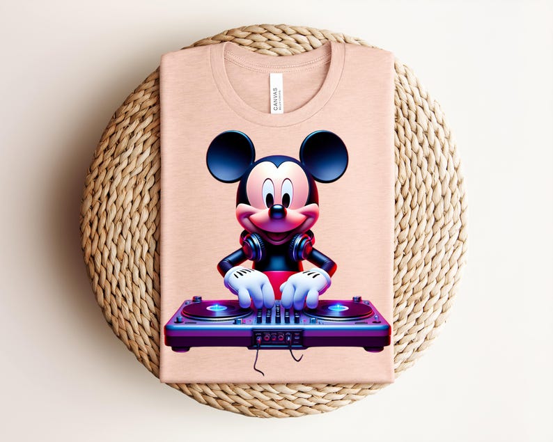 May include: A light peach-colored t-shirt featuring a graphic of Mickey Mouse as a DJ. Mickey is wearing headphones and has his hands on a purple and blue turntable. The shirt is folded on a woven, circular placemat.