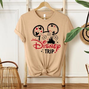 May include: Tan t-shirt featuring a black and red graphic design. The design includes Mickey Mouse ears, fireworks, a castle silhouette, and the words "Disney TRIP". The shirt is knotted at the waist and hangs on a wooden hanger.