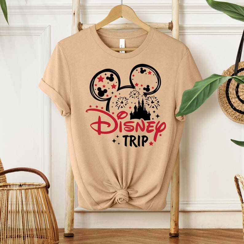May include: Tan t-shirt featuring a black and red graphic design. The design includes Mickey Mouse ears, fireworks, a castle silhouette, and the words "Disney TRIP". The shirt is knotted at the waist and hangs on a wooden hanger.