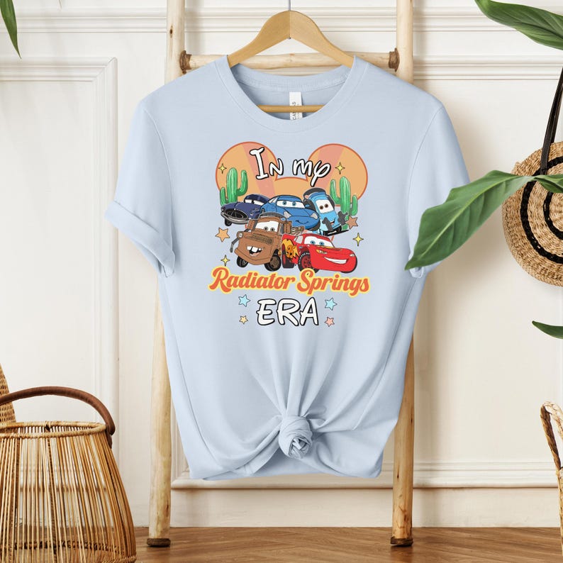 K&ouml;nnte beinhalten: Hellblaues T-Shirt mit einer Grafik von Zeichentrickautos und dem Text "In my Radiator Springs ERA". Das Design umfasst Kakteen und Sterne. Das T-Shirt h&auml;ngt an einem Holzgestell.