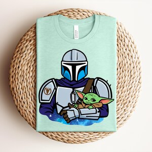 Puede incluir: Camiseta verde claro con una ilustraci&oacute;n de dibujos animados de un personaje mandaloriano sosteniendo a un alien&iacute;gena verde. El mandaloriano lleva una armadura plateada con detalles azules. El alien&iacute;gena est&aacute; envuelto en una manta marr&oacute;n.