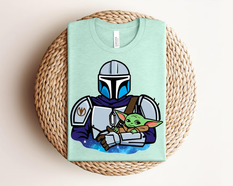 Puede incluir: Camiseta verde claro con una ilustraci&oacute;n de dibujos animados de un personaje mandaloriano sosteniendo a un alien&iacute;gena verde. El mandaloriano lleva una armadura plateada con detalles azules. El alien&iacute;gena est&aacute; envuelto en una manta marr&oacute;n.