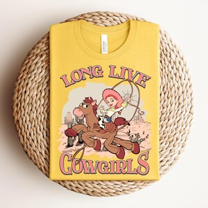 May include: A mustard yellow t-shirt featuring a graphic print of a cartoon cowgirl riding a brown horse in a desert landscape. The text "LONG LIVE COWGIRLS" is printed in pink with a dark outline above and below the central illustration.