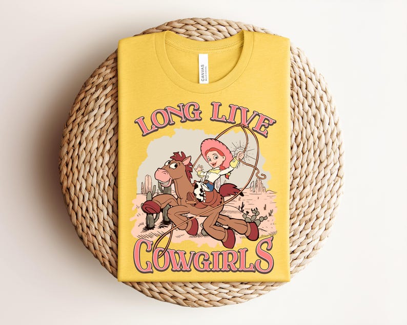 May include: A mustard yellow t-shirt featuring a graphic print of a cartoon cowgirl riding a brown horse in a desert landscape. The text "LONG LIVE COWGIRLS" is printed in pink with a dark outline above and below the central illustration.