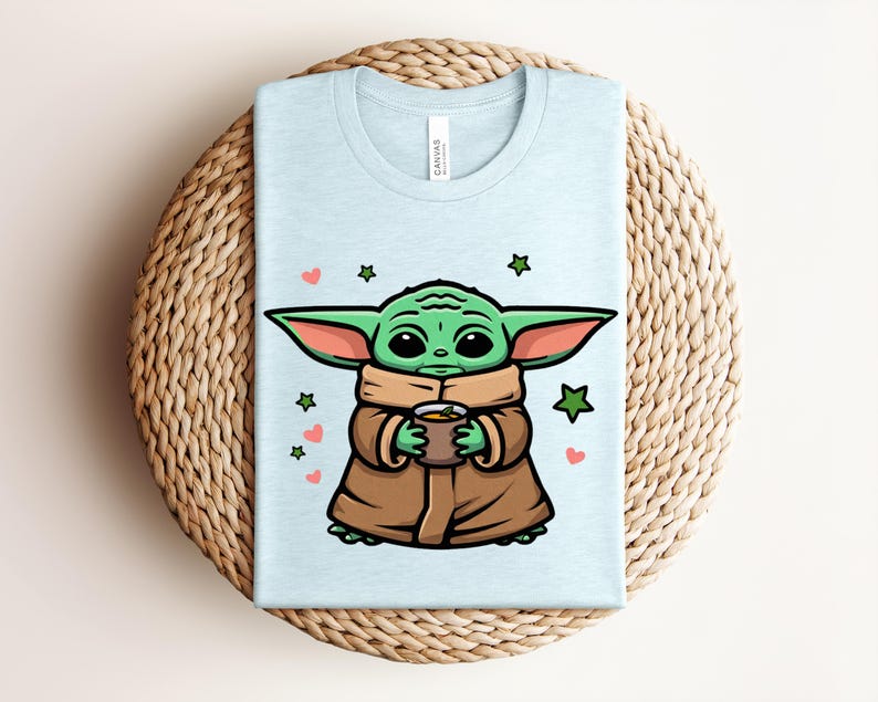May include: Light blue t-shirt featuring a cartoon character holding a cup. The character has green skin, large ears, and is wearing a brown cloak. The design includes green stars and pink hearts. The shirt is folded on a woven surface.
