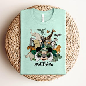 Puede incluir: Una camiseta de color verde menta claro con un estampado gr&aacute;fico de varios animales y personajes de dibujos animados con equipo de safari. El dise&ntilde;o incluye binoculares, hojas y el texto Disney's ANIMAL KINGDOM impreso en letras negras.