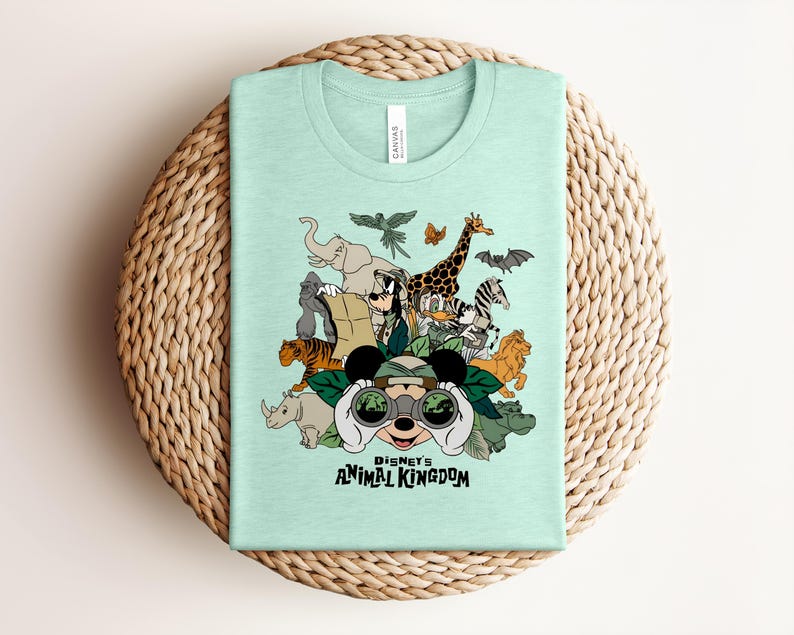 Puede incluir: Una camiseta de color verde menta claro con un estampado gr&aacute;fico de varios animales y personajes de dibujos animados con equipo de safari. El dise&ntilde;o incluye binoculares, hojas y el texto Disney's ANIMAL KINGDOM impreso en letras negras.