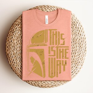 May include: A peach-colored crew neck t-shirt featuring a distressed gold graphic of a Mandalorian helmet on the left and the text THIS IS THE WAY in a bold, stylized font on the right. The shirt has a white brand label at the inner neckline.
