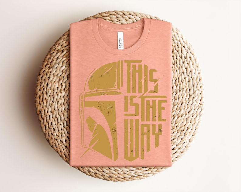 May include: A peach-colored crew neck t-shirt featuring a distressed gold graphic of a Mandalorian helmet on the left and the text THIS IS THE WAY in a bold, stylized font on the right. The shirt has a white brand label at the inner neckline.
