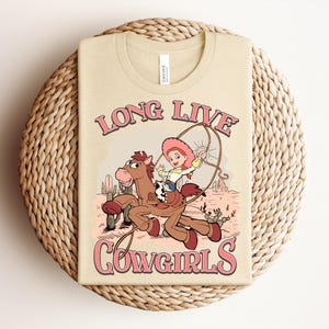 May include: A cream-colored t-shirt featuring a graphic of a cartoon cowgirl riding a brown horse while holding a lasso. The text "LONG LIVE COWGIRLS" is printed in pink with a dark outline above and below the illustration in a desert setting.