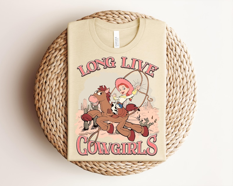 May include: A cream-colored t-shirt featuring a graphic of a cartoon cowgirl riding a brown horse while holding a lasso. The text "LONG LIVE COWGIRLS" is printed in pink with a dark outline above and below the illustration in a desert setting.