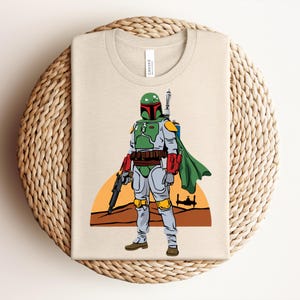 May include: Beige t-shirt featuring a colorful illustration of a character from a science fiction film. The character is depicted in a green and gray suit with a green helmet, red accents, and a cape. The design includes a blaster and a desert landscape.