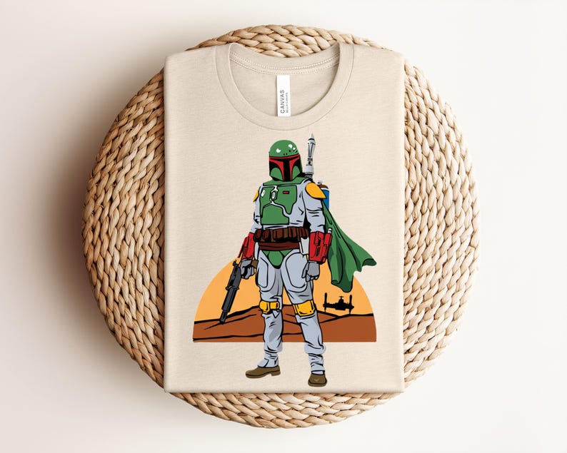 May include: Beige t-shirt featuring a colorful illustration of a character from a science fiction film. The character is depicted in a green and gray suit with a green helmet, red accents, and a cape. The design includes a blaster and a desert landscape.