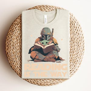 May include: A light beige t-shirt featuring a graphic of a helmeted armored figure sitting cross-legged and reading a book with a small green creature. The text "READING IS THE WAY" is printed in large, light orange letters at the bottom of the shirt.