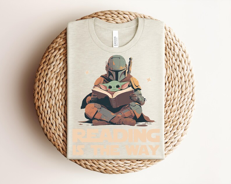 May include: A light beige t-shirt featuring a graphic of a helmeted armored figure sitting cross-legged and reading a book with a small green creature. The text "READING IS THE WAY" is printed in large, light orange letters at the bottom of the shirt.