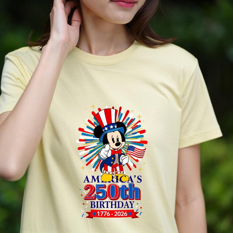 May include: A light yellow t-shirt featuring a graphic of Mickey Mouse dressed in patriotic attire, holding an American flag. The graphic includes the text "AMERICA'S 250th BIRTHDAY 1776-2026" in red, white, and blue.