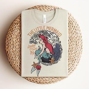 May include: A light beige t-shirt featuring a graphic of a red-haired mermaid with a teal tail sitting on a rock in front of a large wave. The text reads THE LITTLE MERMAID, Under the Sea, and '89. Two orange starfish and a pink seashell are visible.