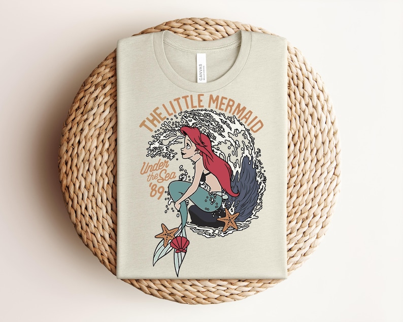 May include: A light beige t-shirt featuring a graphic of a red-haired mermaid with a teal tail sitting on a rock in front of a large wave. The text reads THE LITTLE MERMAID, Under the Sea, and '89. Two orange starfish and a pink seashell are visible.