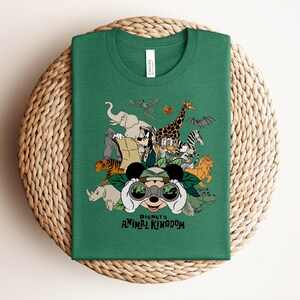 Puede incluir: Una camiseta de cuello redondo color verde bosque con un estampado gr&aacute;fico de Mickey Mouse mirando a trav&eacute;s de binoculares rodeado de varios animales, incluyendo un elefante, jirafa, tigre y cebra, con el texto Disney's Animal Kingdom debajo.