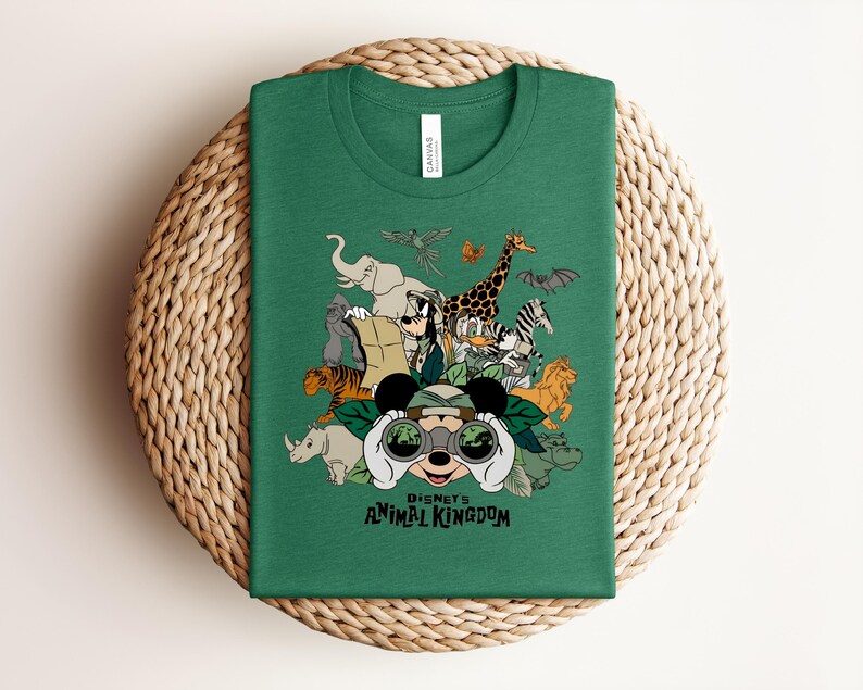 Puede incluir: Una camiseta de cuello redondo color verde bosque con un estampado gr&aacute;fico de Mickey Mouse mirando a trav&eacute;s de binoculares rodeado de varios animales, incluyendo un elefante, jirafa, tigre y cebra, con el texto Disney's Animal Kingdom debajo.