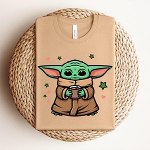 May include: A tan t-shirt featuring a cartoon illustration of a green alien character holding a cup. The character is wearing a brown cloak, with green ears and large black eyes. Pink hearts and green stars surround the character.