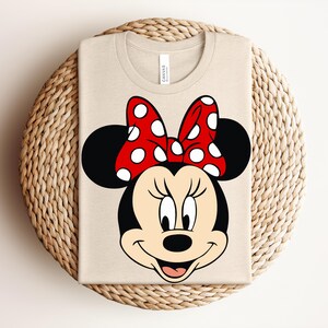 May include: A beige t-shirt featuring a large graphic of Minnie Mouse's face. The design includes black ears, a red bow with white polka dots, and a smiling face. The shirt is folded on a woven, circular surface.