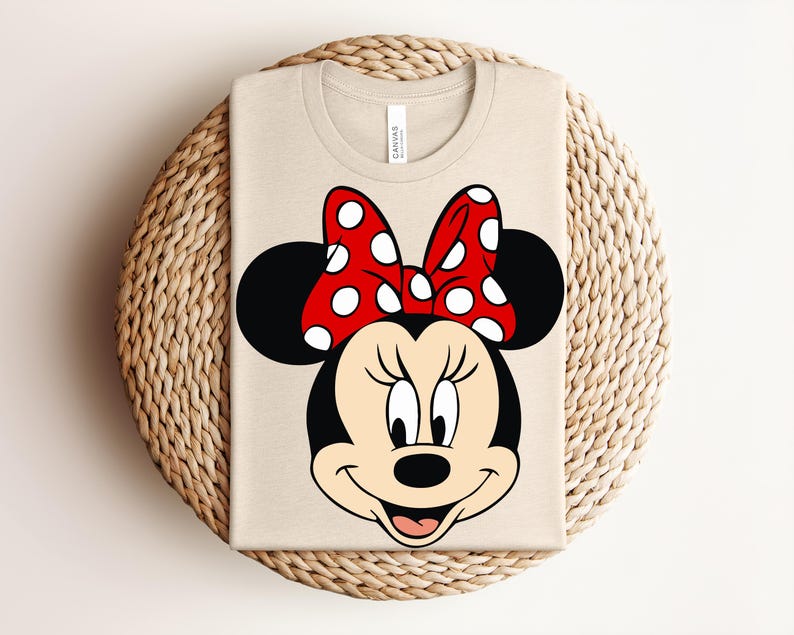 May include: A beige t-shirt featuring a large graphic of Minnie Mouse's face. The design includes black ears, a red bow with white polka dots, and a smiling face. The shirt is folded on a woven, circular surface.