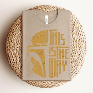 May include: A light tan crew neck t-shirt featuring a distressed mustard yellow graphic of a helmet silhouette on the left and the text "THIS IS THE WAY" in a bold, blocky, distressed font on the right side of the chest area.