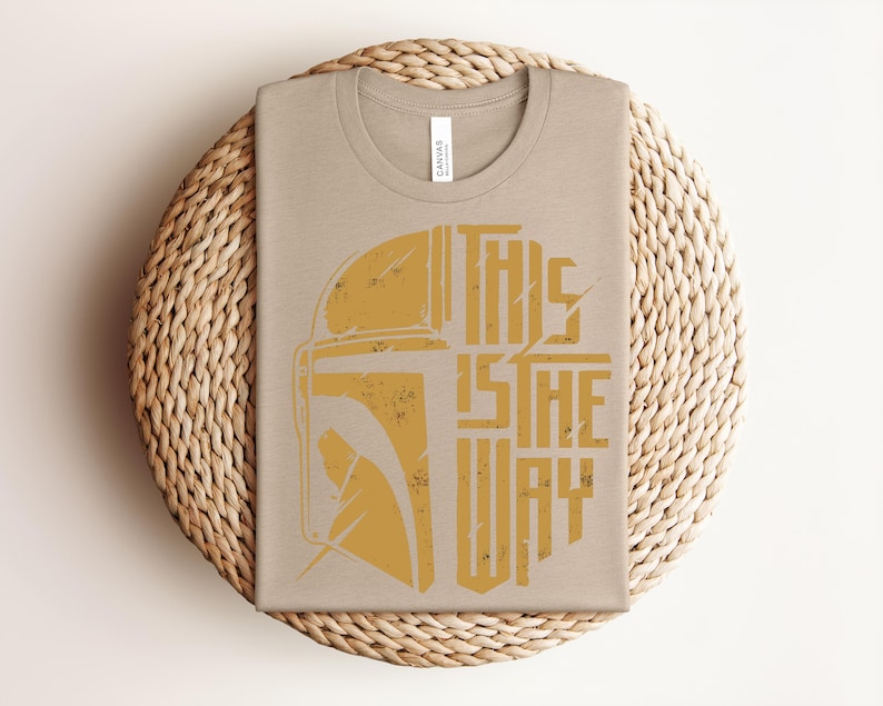 May include: A light tan crew neck t-shirt featuring a distressed mustard yellow graphic of a helmet silhouette on the left and the text "THIS IS THE WAY" in a bold, blocky, distressed font on the right side of the chest area.