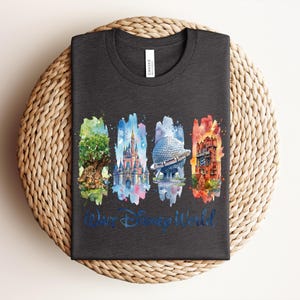May include: Dark gray t-shirt featuring a watercolor design of four iconic Disney World landmarks: the Tree of Life, Cinderella Castle, Spaceship Earth, and the Tower of Terror. The words "Walt Disney World" are written in blue script below the images. The shirt is folded on a woven mat.