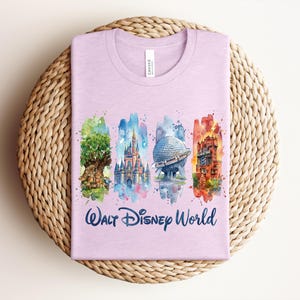 May include: A light purple t-shirt featuring watercolor illustrations of iconic Walt Disney World landmarks. The shirt includes images of the Tree of Life, Cinderella's Castle, Spaceship Earth, and the Tower of Terror, with "Walt Disney World" written below.