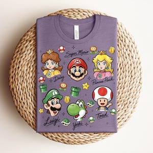 May include: A purple t-shirt featuring cartoon illustrations of Super Mario characters, including Mario, Luigi, Princess Peach, Daisy, Yoshi, and Toad. The words "Super Mario" and character names are written on the shirt.