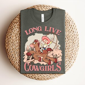 May include: A dark gray t-shirt featuring a graphic of a cartoon cowgirl riding a brown horse in a desert landscape with cacti. The text "LONG LIVE COWGIRLS" is printed in pink above and below the illustration. The shirt has a crew neckline.