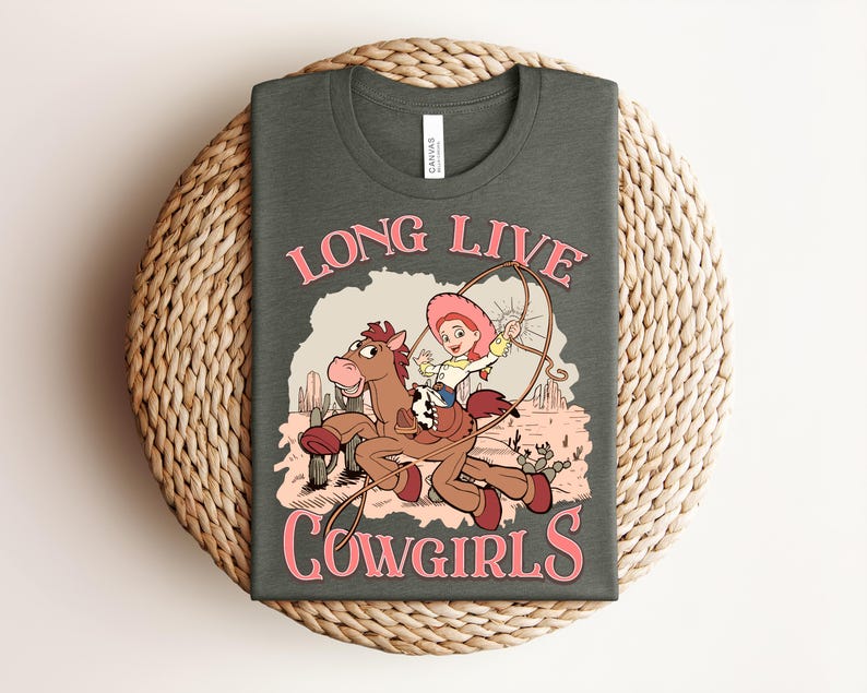 May include: A dark gray t-shirt featuring a graphic of a cartoon cowgirl riding a brown horse in a desert landscape with cacti. The text "LONG LIVE COWGIRLS" is printed in pink above and below the illustration. The shirt has a crew neckline.
