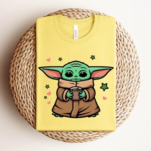 May include: Yellow t-shirt featuring a cartoon illustration of a green alien character holding a cup. The character is wearing a brown cloak and has large ears. Pink hearts and green stars surround the character.