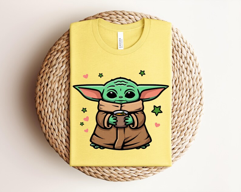 May include: Yellow t-shirt featuring a cartoon illustration of a green alien character holding a cup. The character is wearing a brown cloak and has large ears. Pink hearts and green stars surround the character.