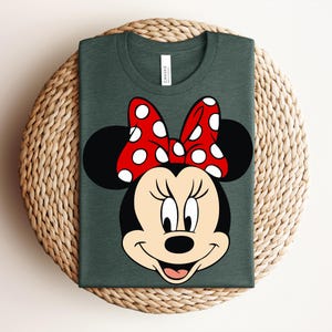May include: Dark green t-shirt featuring a graphic of Minnie Mouse's face. The design includes a red bow with white polka dots, black ears, and a smiling face. The shirt is folded on a woven surface.