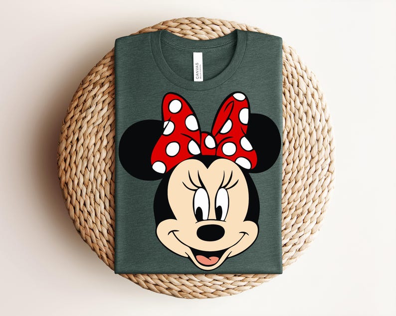 May include: Dark green t-shirt featuring a graphic of Minnie Mouse's face. The design includes a red bow with white polka dots, black ears, and a smiling face. The shirt is folded on a woven surface.