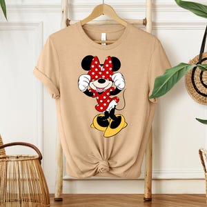 May include: Tan-colored t-shirt featuring a Minnie Mouse graphic. The graphic shows Minnie Mouse in a red dress with white polka dots, a matching bow, and yellow shoes. The shirt is hanging on a wooden hanger.