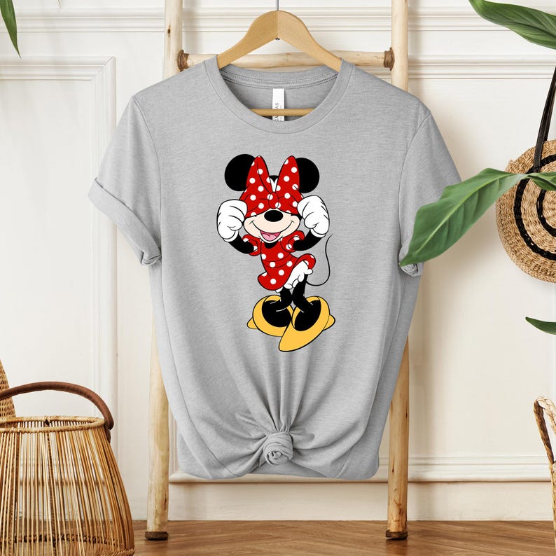 May include: A gray t-shirt featuring a Minnie Mouse graphic. Minnie is depicted in her classic outfit: a red dress with white polka dots, a matching bow, and yellow shoes. The shirt is knotted at the waist and hangs on a wooden hanger.