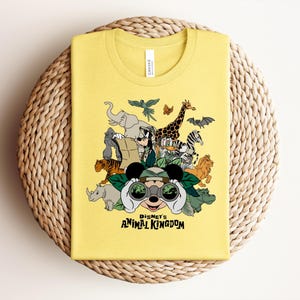 Puede incluir: Una camiseta amarilla con un estampado gr&aacute;fico de varios animales y personajes de dibujos animados con binoculares. El texto "DISNEY'S ANIMAL KINGDOM" est&aacute; impreso en negro debajo de la ilustraci&oacute;n. La camiseta tiene cuello redondo.