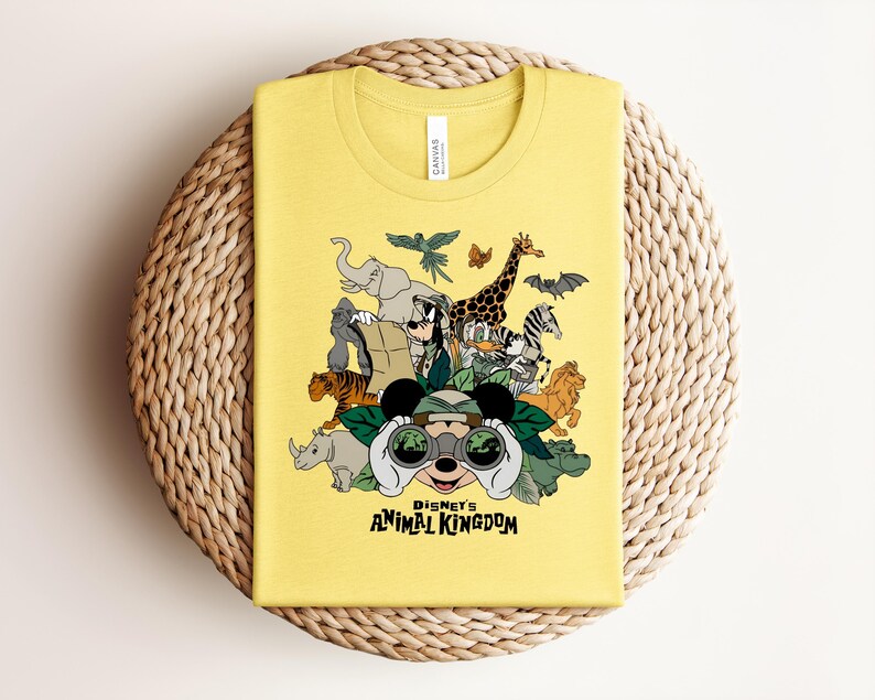 Puede incluir: Una camiseta amarilla con un estampado gr&aacute;fico de varios animales y personajes de dibujos animados con binoculares. El texto "DISNEY'S ANIMAL KINGDOM" est&aacute; impreso en negro debajo de la ilustraci&oacute;n. La camiseta tiene cuello redondo.