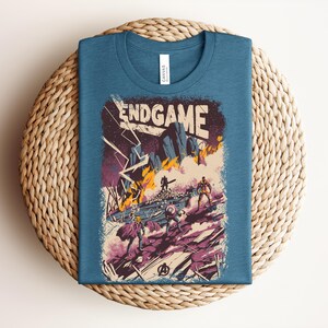 Pu&ograve; includere: Una t-shirt blu a girocollo con una stampa grafica che riporta il testo ENDGAME. L'illustrazione raffigura una scena di battaglia con figure stilizzate, fulmini e fuoco in tonalit&agrave; viola, arancione e bianco, con un logo Avengers in basso.