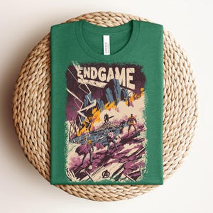 Pu&ograve; includere: Una t-shirt verde bosco con una stampa grafica che riporta il testo ENDGAME in alto. L'illustrazione raffigura una scena di battaglia con diverse figure, terreno roccioso, fulmini e fuoco nelle tonalit&agrave; del viola, arancione e blu.